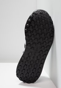 Musta kuminen pohja Adidas-kengästä, jossa on kuusikulmainen kuviointi, asetettuna pystyasentoon valkoista seinää ja pintaa vasten.