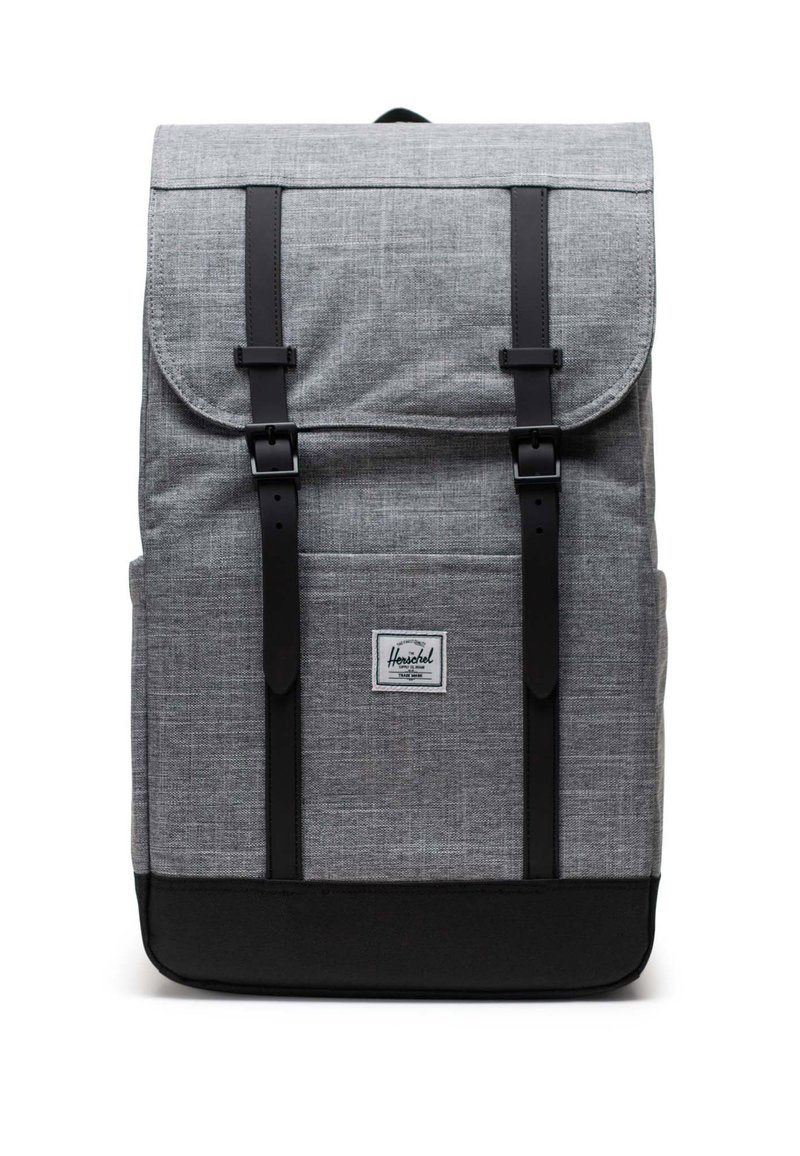 Mochila de tela gris con base negra, dos correas de hebilla negra, cierre con solapa y parche de etiqueta en la parte frontal sobre un fondo blanco.