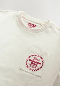 Camiseta color crema de corte relajado, con un gráfico vintage rojo en el bolsillo izquierdo. Textura de tela de algodón suave.
