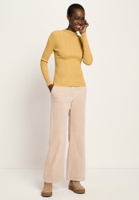 Lächelnde Frau in senfgelbem Rippstrickpullover, beigen weiten Hosen und braunen Plateauschuhen, die mit einer Hand in der Tasche steht.