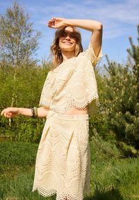 Femme souriante portant une tenue deux-pièces en dentelle crème, debout à l'extérieur avec un bras levé, entourée d'arbres verts et d'un ciel bleu.