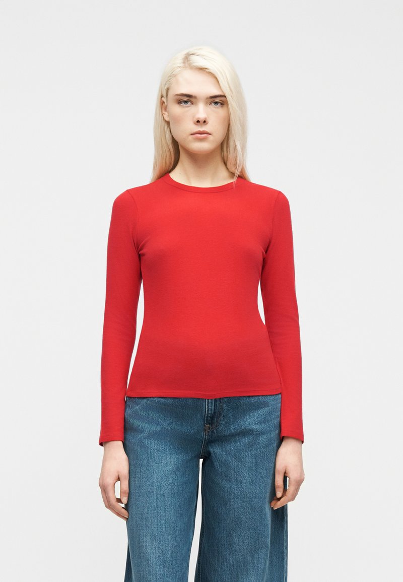 Γυναίκα με μακριά ξανθά μαλλιά που φοράει εφαρμοστή κόκκινη μπλούζα με μακριά μανίκια και άνετο μπλε τζιν, στέκεται μπροστά σε ένα απλό φωτεινό φόντο.