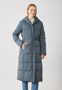 Νεαρή γυναίκα που χαμογελά, φορώντας έναν ραμμένο, μακρύ μπλε καπιτονέ μπουφάν με κουκούλα, στέκεται με το ένα χέρι στην τσέπη σε ένα απλό φόντο.