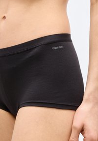 Κοντινή εικόνα στη μέση και το πάνω μέρος του μηρού ενός ατόμου που φοράει στενά εσώρουχα της Organic Basics σε μαύρο χρώμα με ορατό λογότυπο της μάρκας.