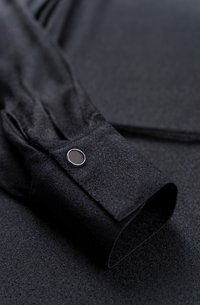 Schwarze Stoffhemdmanschette mit glatter Textur, Knopfdetail mit metallischem Akzent, aufgerollter Stil vor einem dunklen Hintergrund.