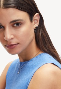 Boucles d'oreilles noires avec un accent floral et un pendentif circulaire. Le sujet porte un haut sans manches bleu côtelé et un délicat collier en argent.