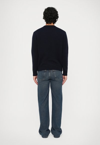 Un uomo con capelli ricci e scuri sta in piedi di spalle indossando un maglione nero e jeans larghi blu contro uno sfondo bianco uniforme.