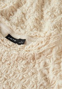 Haut texturé de couleur crème avec un motif en dentelle. Doté d'un col rond et d'une étiquette portant "LMTD" avec une étiquette de taille. Tissu doux et léger.