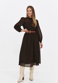 Czarna sukienka z długim rękawem w pomarańczowy i brązowy wzór w kropki, z zaznaczoną talią i brązowym paskiem, zestawiona z beżowymi kozakami do kolan.