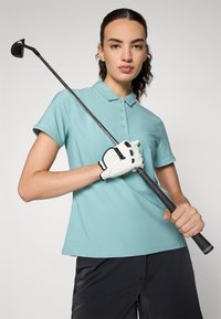 Světle modrá polo tričko s límečkem, krátkými rukávy a hladkým povrchem, v kombinaci s bílou golfovou rukavicí, držící golfovou hůl.