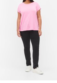 Lyserød T-shirt med korte ærmer og op-rullede ærmer, parret med sorte slim-fit jeans. Model iført hvide sneakers, stående mod en ensfarvet baggrund.