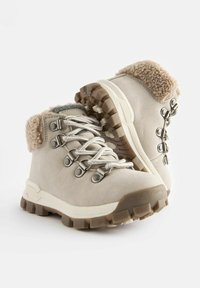 Beige mockaskor för vandring med en fluffig beige krage, grå foder, metallöglor och slitstarka bruna gummisulor. Har snörning och krokar.