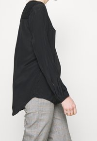 Blusa nera a maniche lunghe con vestibilità comoda, texture satinata, polsini bottati e orlo asimmetrico, abbinata a pantaloni grigi a quadri.