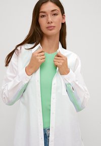 White button-up shirt with long sleeves featuring a green and blue striped accent. Worn over a light green ribbed top.