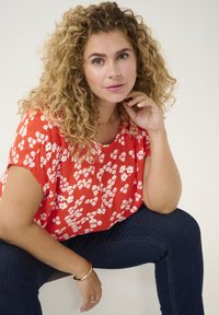 Rød blomsterbluse med korte ærmer, blød tekstur og en afslappet pasform, parret med mørkeblå jeans. Enkel guldarmbånd på håndleddet.