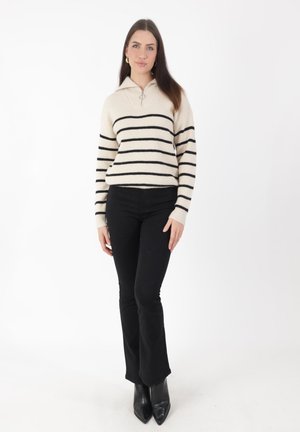 Pull en tricot blanc cassé avec des rayures horizontales noires, col zippé, associé à un pantalon noir évasé et des bottes noires à bout pointu.