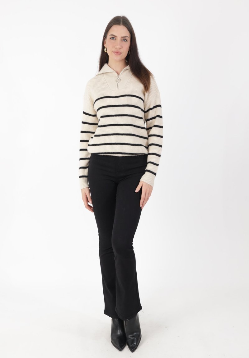 Gestreifter Pullover in Creme mit schwarzen horizontalen Streifen, mit Reißverschlusskragen, kombiniert mit schwarzen, ausgestellten Hosen und schwarzen spitzen Stiefeln.