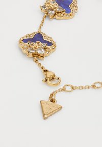 Gouden armband met paarse cabochons, kristalaccenten en een driehoekige logocharme. Inclusief een sluiting en kettinglinken.