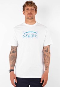 Camiseta de algodón blanca con un logo "OXBOW" en azul y púrpura con acentos de mariposa. Manga corta, corte estándar, textura suave.
