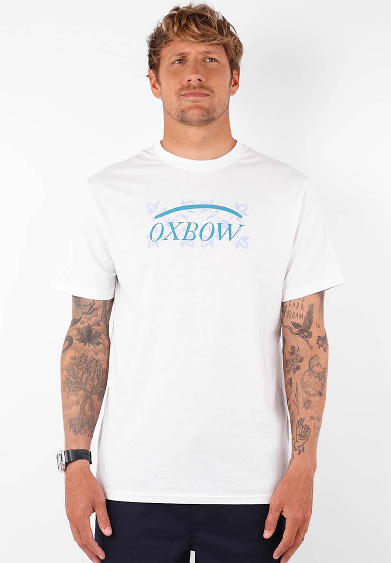 Camiseta de algodón blanca con un logo "OXBOW" en azul y púrpura con acentos de mariposa. Manga corta, corte estándar, textura suave.