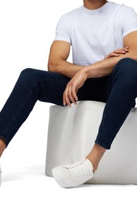 Homme portant un t-shirt blanc, un jean bleu foncé et des baskets blanches assis, les bras croisés, sur un bloc gris sur fond blanc.
