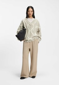 Suéter de punto de color gris claro con patrón y cuello en uve, combinado con pantalones beige de pierna ancha. Sosteniendo un bolso negro acolchado. Zapatos planos negros.