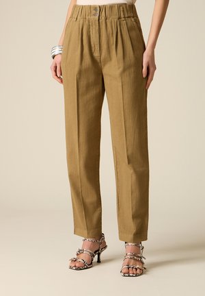 Kobieta w brązowych, wysokowzrostowych spodniach zwężanych z plisami oraz sandałach na obcasie z paskami w wężowy wzór, stojąca na tle o neutralnym kolorze.