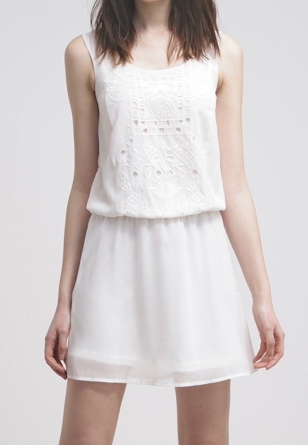 Femme portant une robe blanche sans manches avec des motifs géométriques et floraux brodés sur le corsage, sur un fond uni.
