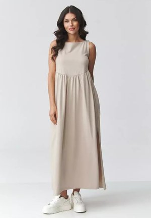 Kobieta o długich, ciemnych włosach, ubrana w bezową, bezrękawnikową sukienkę maxi i białe sneakersy na platformie, stojąca na tle jednolitego, jasnego tła.