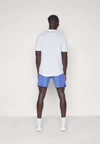 Weißes sportliches T-Shirt mit kurzen Ärmeln, kombiniert mit blauen Shorts. Das Modell steht mit dem Rücken zur Kamera, trägt weiße Sneaker und Knöchelsocken.
