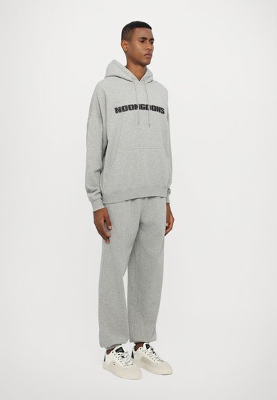 Felpa con cappuccio grigia con il logo "NOON GOONS", abbinata a pantaloni della tuta coordinati e sneakers bianche. Il tessuto sembra morbido e con una texture interessante.