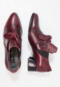 Chaussures en cuir bordeaux avec bout arrondi, nœud décoratif, panneaux latéraux élastiques et talon empilé. Texture lisse, finition soignée.