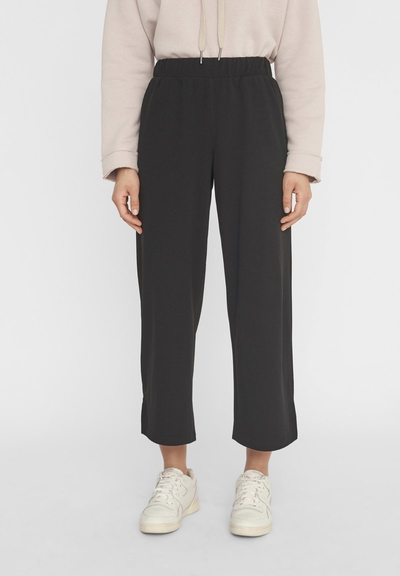 Personne portant un pantalon large noir raccourci, un haut beige à manches longues et des baskets blanches, debout devant un fond clair uni.
