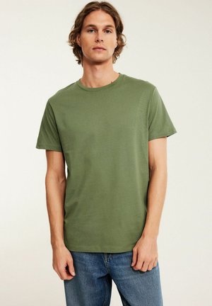 Mann mit mittelbraunen Haaren mittlerer Länge, der ein olivgrünes kurzärmeliges T-Shirt und blaue Jeans trägt und vor einem schlichten Hintergrund steht.