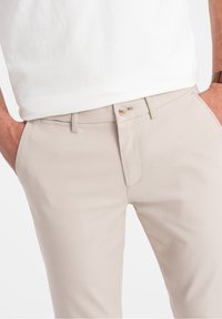 Mężczyzna w beżowych spodniach i białej koszuli z rękami w kieszeniach, pokazujący zapięcie na guzik i pętle na pasek.