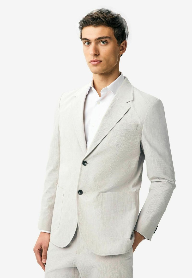 Világosszürke blézer, texturált anyagból, slim fit fazonban, egysoros kialakítással, két elülső zsebbel és fekete gombokkal. Alatta fehér ing.