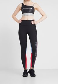 Zwarte sportbeha met zilveren accenten en Puma-logo; gecombineerd met zwarte leggings met blauwe en rode panelen en bedrukte tekst.