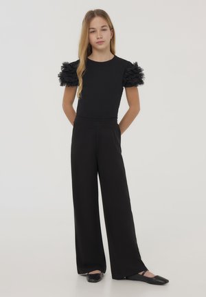 Top nero con maniche in tulle arricciate e pantaloni neri a gamba larga. Design semplice, texture liscia, completato da scarpe nere basse.