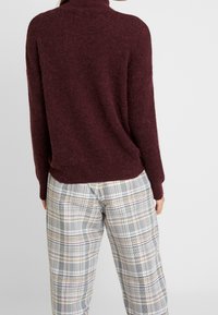 Suéter de cuello alto burdeos de textura suave, combinado con pantalones de cuadros grises con finas líneas amarillas, rojas y negras.