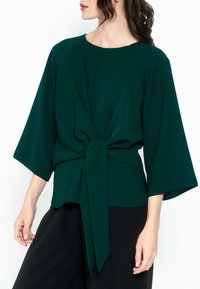 Top verde a maniche corte con scollo rotondo, dettaglio anteriore annodato e vestibilità rilassata, realizzato in tessuto morbido con una leggera lucentezza.