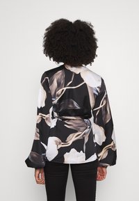Blouse noire à manches longues avec un motif floral comportant des fleurs blanches et beiges, taille cintrée et texture de tissu lisse.