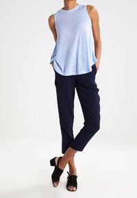 Donna con top senza maniche blu chiaro, pantaloni scuri blu a vita alta, e sandali a pianta aperta neri con tacco, in piedi con le mani in tasca.