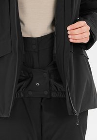 Svart ytterklädsel med en zipjacka och byxor med tryckknappar. Slät textur med elastisk midja och manschetter för en säker passform.