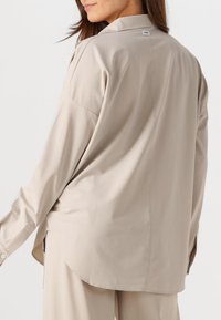 Beige Langarmhemd mit entspannter Passform, Kragen und seitlichen Schlitz. Glatte Textur und minimale Nähdetails auf dem Rücken.