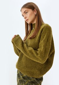 Oliwkowy sweter dzianinowy z wysokim kołnierzem, luźnym krojem i długimi rękawami. Tekstura jest miękka, z wyraźnym, prążkowanym wzorem.