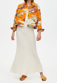 Džínová bunda s kamuflážním vzorem v oranžové a zelené barvě přes bílou úpletovou maxi šaty se strukturovaným zigzagovým vzorem a žlutými sandály na slidě.