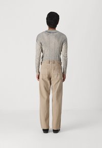 Person sedd bakifrån i en ribbade långärmad tröja, beige högmidjade byxor med bakfickor och svarta skor, stående på ett vitt golv.