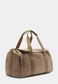 Borsa duffel beige realizzata in tessuto liscio, con manici doppi, chiusura con zip e dettagli in nastro. Forma arrotondata con un design minimale.