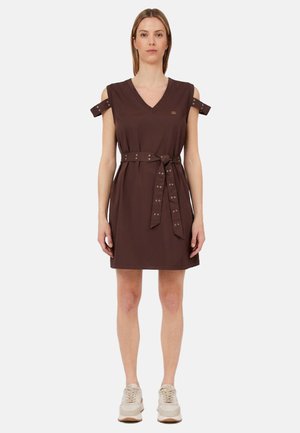 Donna che indossa un vestito marrone senza maniche con scollo a V, spalline e cintura annodata, abbinato a sneakers beige, in piedi davanti a uno sfondo semplice.