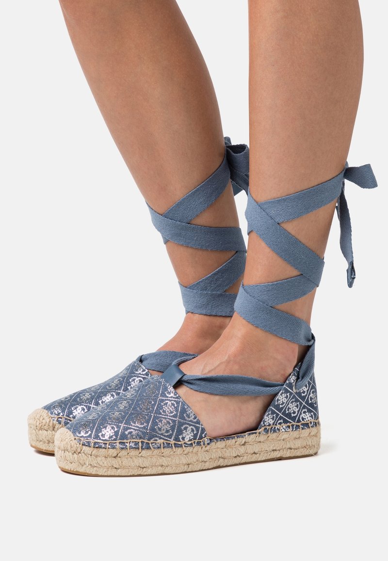 Pieds portant des espadrilles à motifs bleus avec des rubans croisés autour de la cheville noués au-dessus des chevilles sur un fond neutre.